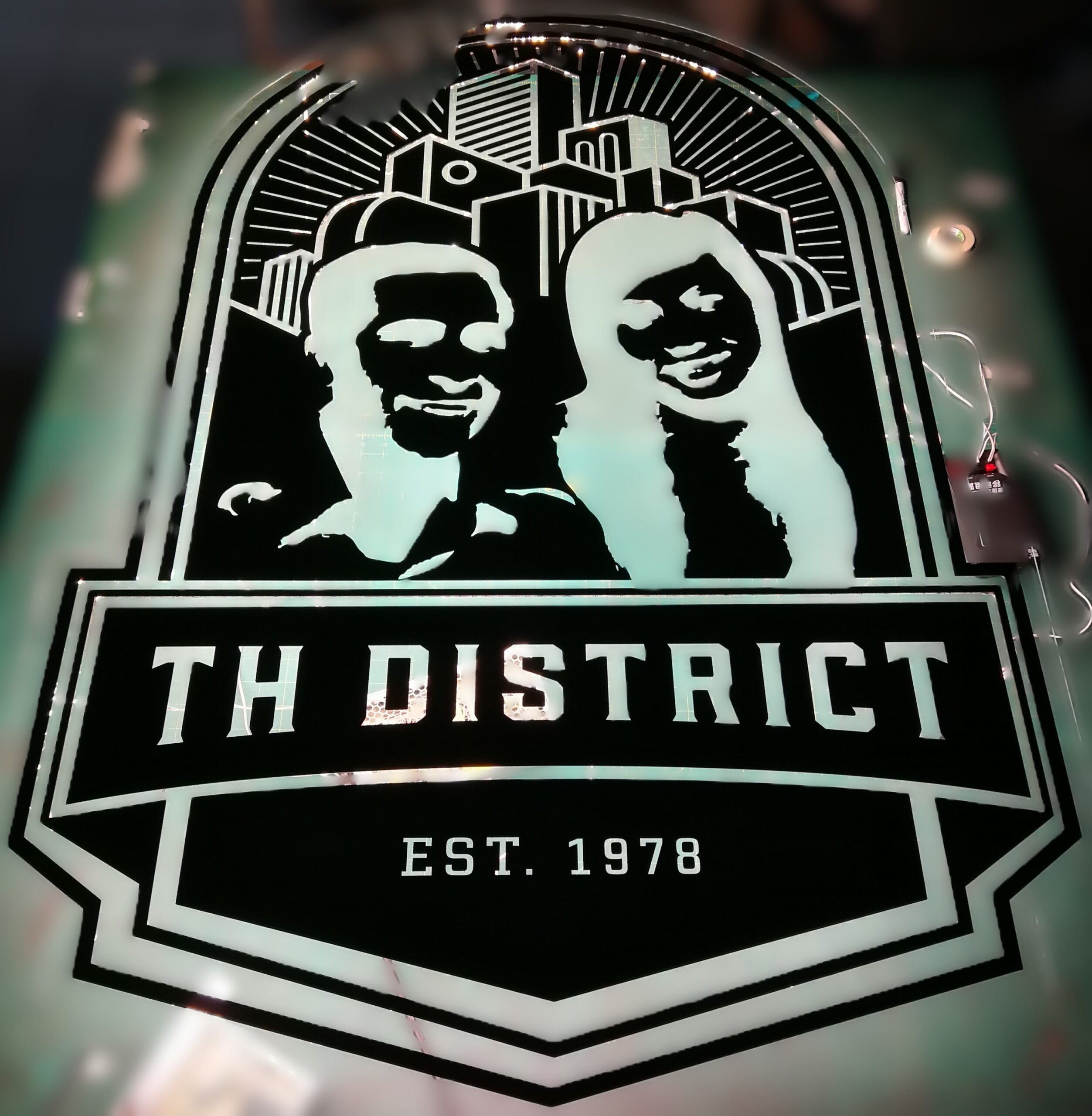 ป้ายไฟส่องหลัง TH DISTRICT EST. 1978 รูปคู่รักหน้าตึกเมืองทรงโล่