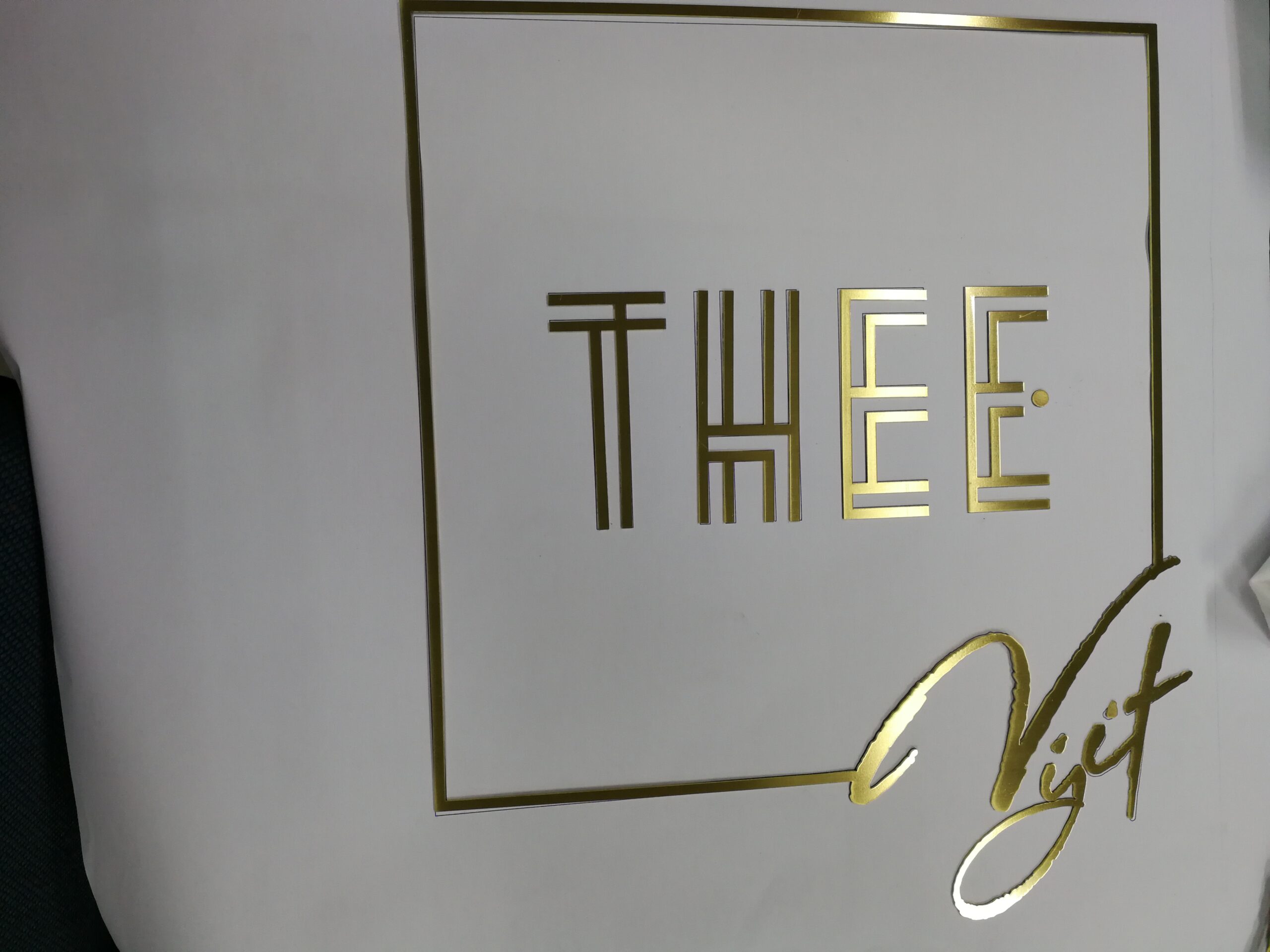 ป้ายสแตนเลสสีทองตัวอักษร THEE สไตล์ Art Deco พร้อมลายเซ็น Viet ติดผนังขาว