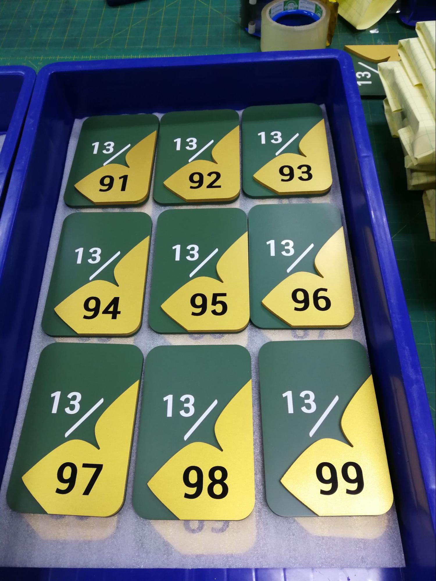 ป้ายเลขที่ห้องคอนโดสีเขียวทอง ชั้น 13 ห้อง 91-99 พร้อมโลโก้ใบไม้ วางเรียงในถาดที่โรงงาน