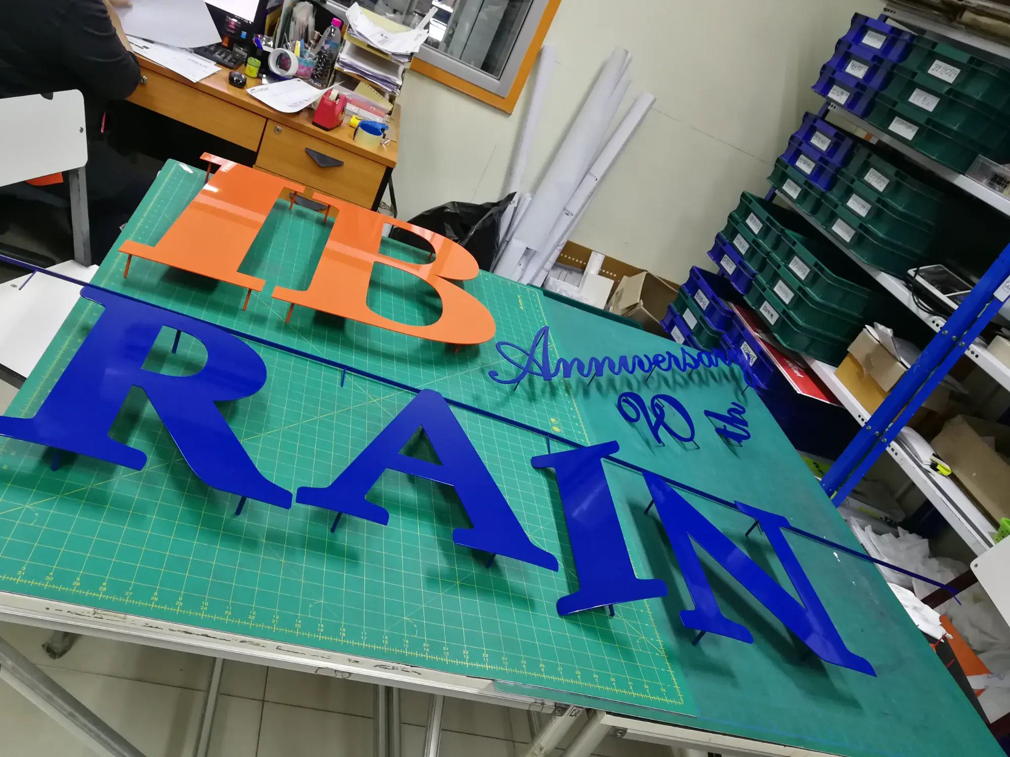 ตัวอักษรอะคริลิกสีส้มและน้ำเงิน IB BRAIN Anniversary 90th บนโต๊ะตัด