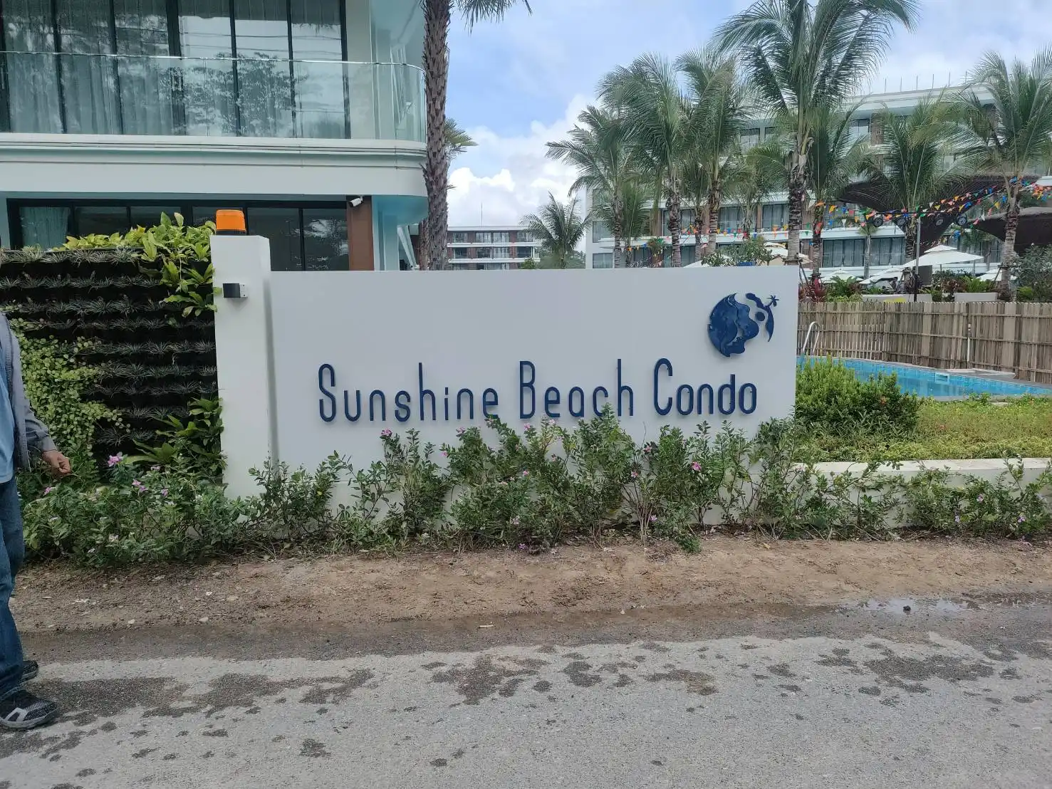 ป้ายชื่อคอนโด Sunshine Beach Condo ตัวอักษรสแตนเลสสีน้ำเงินพร้อมโลโก้ลูกโลก ติดตั้งบนกำแพงสีขาว