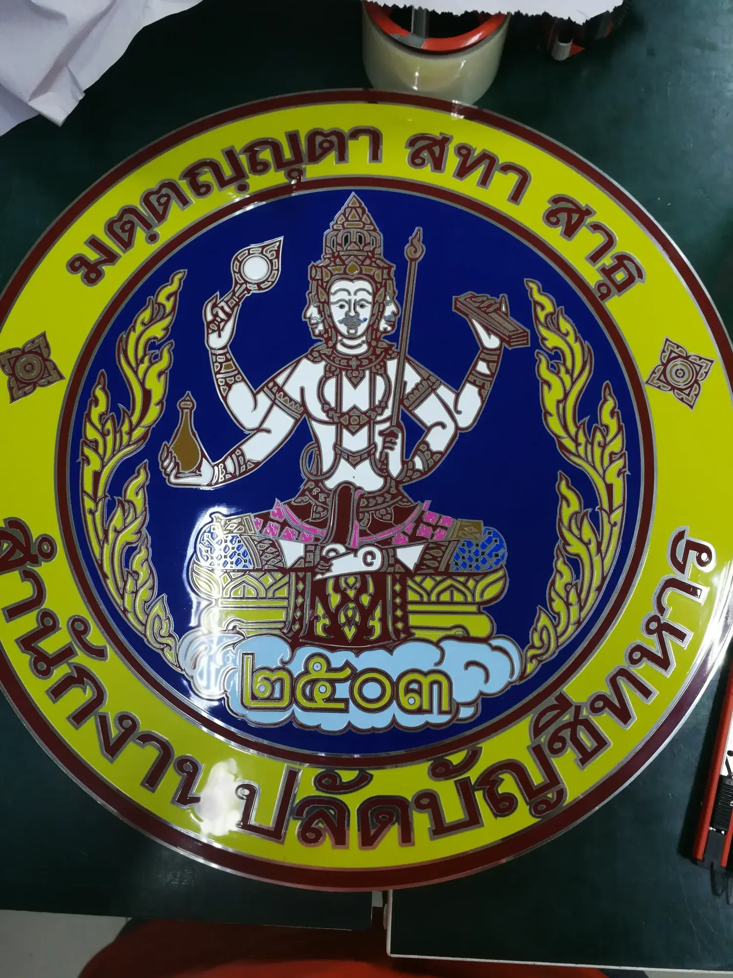ป้ายตราสัญลักษณ์สำนักงานปลัดบัญชีทหาร รูปเทพถือเครื่องมือช่างบนพื้นสีน้ำเงินขอบเหลือง ปี 2503