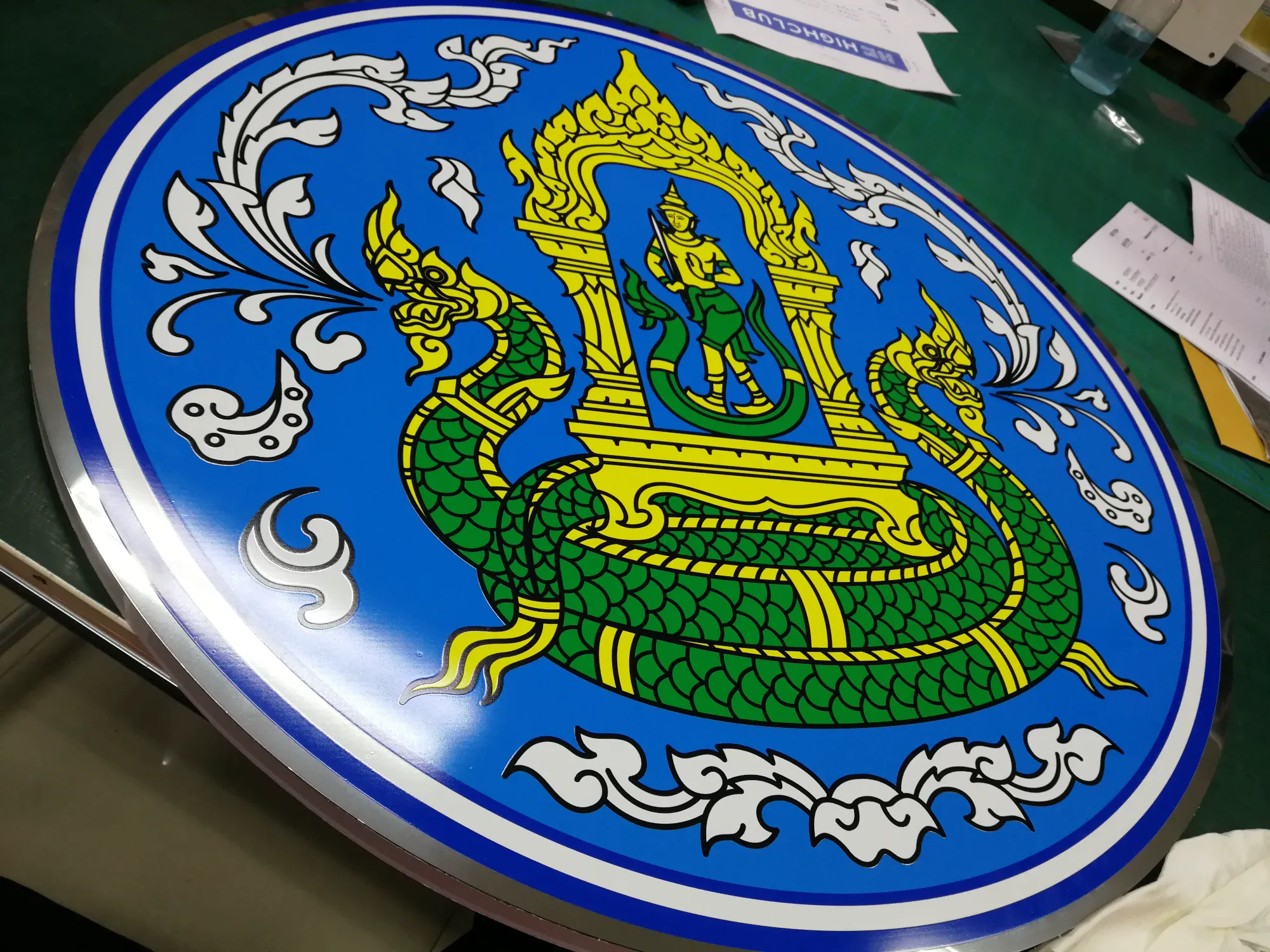 ป้ายตราสัญลักษณ์รูปพญานาคสองตัวสีเขียวล้อมเทพธิดาใต้ซุ้มทองบนพื้นสีน้ำเงิน ขอบสแตนเลส