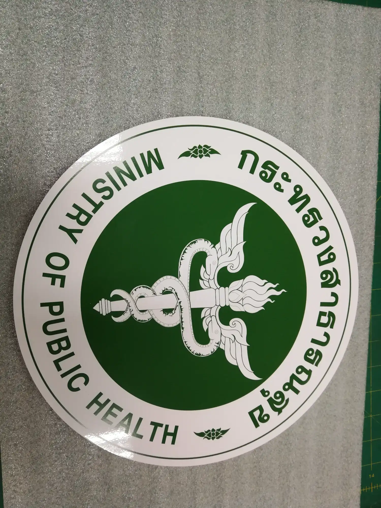 ป้ายตราสัญลักษณ์กระทรวงสาธารณสุข รูปคทางูพันปีกบนพื้นเขียว ข้อความไทยและอังกฤษ