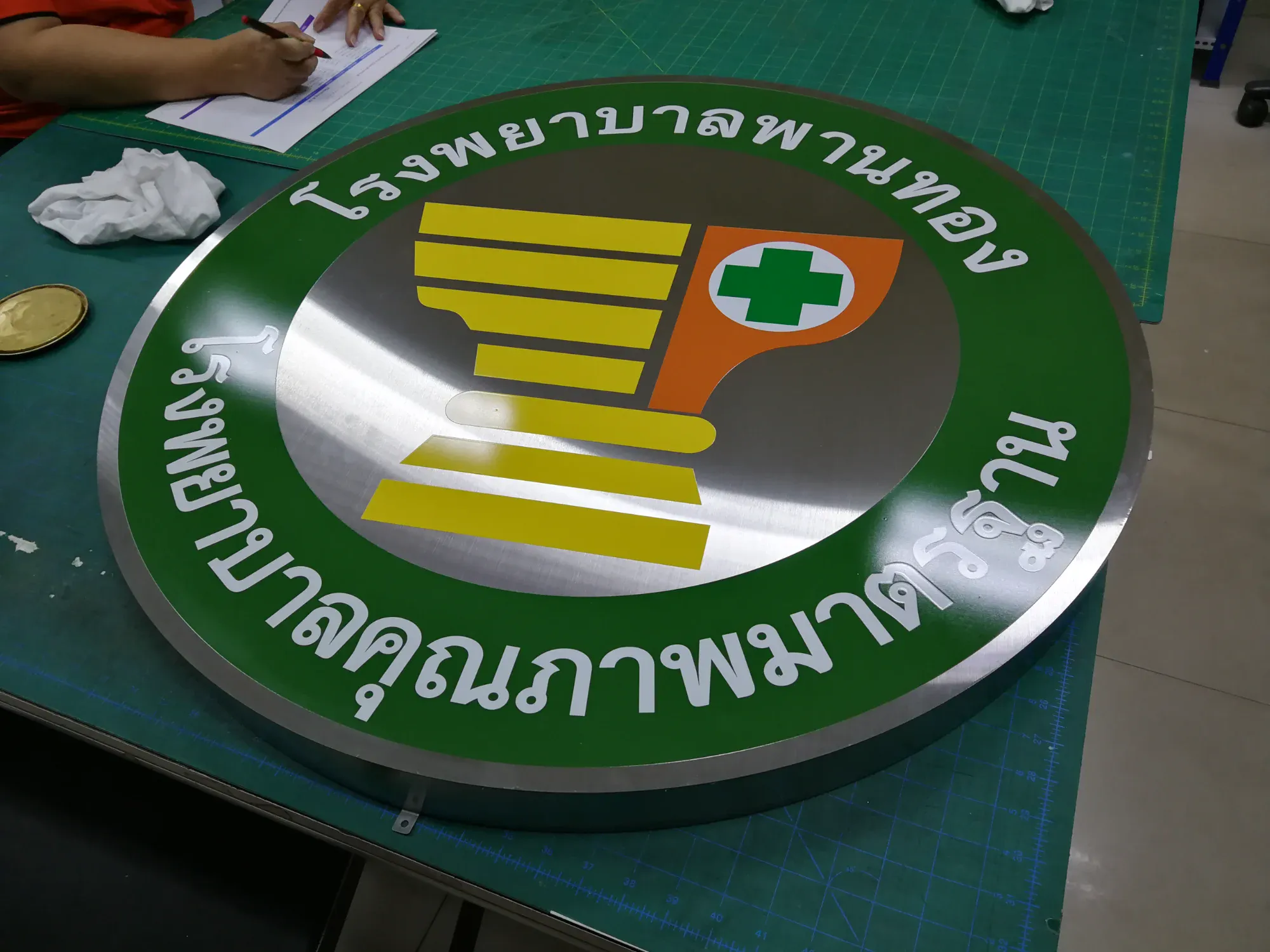 ป้ายตราสัญลักษณ์โรงพยาบาลพานทอง สแตนเลส รูปครกยาสีทองพร้อมกากบาทเขียวบนพื้นเขียว